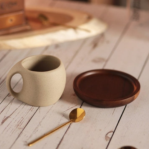 A beige ceramic coffee mug with a gold-colored spoon next to it, also a brown wooden saucer, which is placed on a wooden surface.