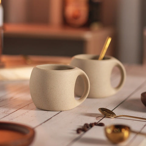 A beige ceramic coffee mug with a gold-colored spoon in it, resting on a brown wooden saucer, which is placed on a wooden surface with coffee beans scattered around. A second mug and some packaging are visible in the background.