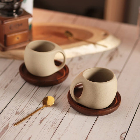 A beige ceramic coffee mug with a gold-colored spoon in it, resting on a brown wooden saucer, which is placed on a wooden surface with coffee beans scattered around. A second mug and some packaging are visible in the background.
