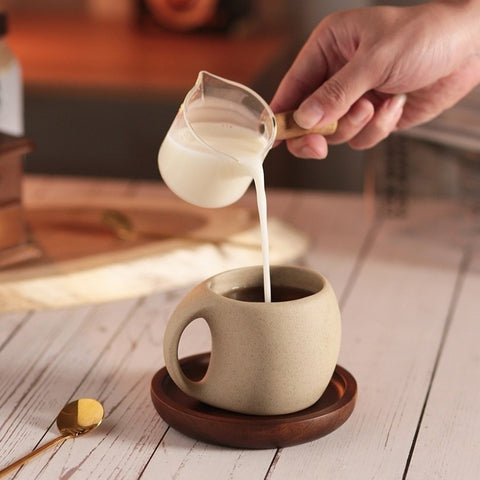 A beige ceramic coffee mug resting on a brown wooden saucer, which is placed on a wooden surface, golden spoon also resting on wooden table near coffee mug, hand pouring milk in the coffee mug with using glass measuring cup.