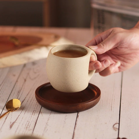 A beige ceramic coffee mug with a gold-colored spoon in it, resting on a brown wooden saucer, which is placed on a wooden surface, hand reaching for a mug.