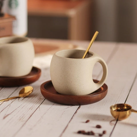 A beige ceramic coffee mug with a gold-colored spoon in it, resting on a brown wooden saucer, which is placed on a wooden surface with coffee beans scattered around. A second mug and some packaging are visible in the background.