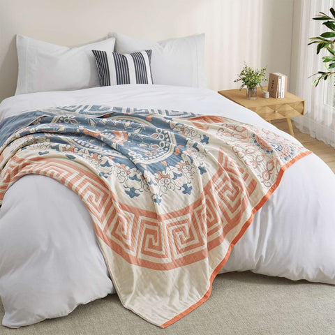 Lightweight cotton throw blanket with orange and blue geometric design draped over a white bed in a cozy natural-light bedroom.
