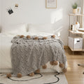 Gray knitted blanket with pom-poms on a bed in a bedroom.
