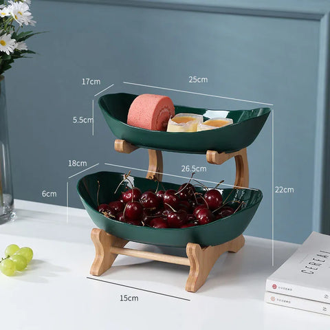 Two-tiered green fruit bowl with wooden stand containing fruits and measurements on a white surface.
