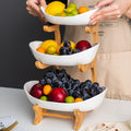 Three-tiered fruit basket with various fruits being added, person wearing a beige apron.