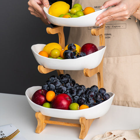 Three-tiered fruit basket with various fruits being added, person wearing a beige apron.