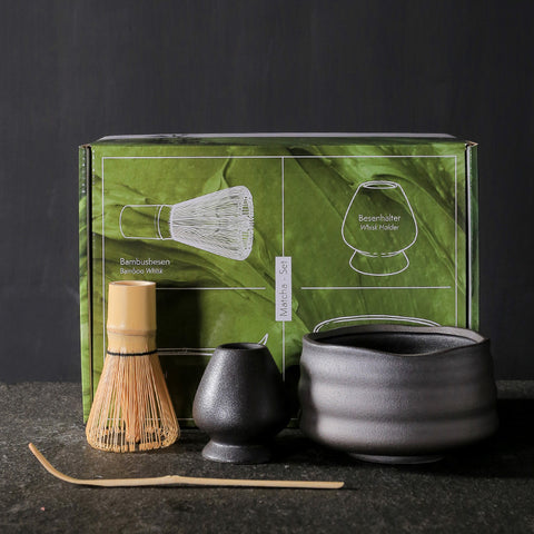 Tea-making set with black teapot, whisk, and holder in front of a green box on a dark background