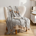 A gray Nordic-inspired knitted throw with a cable knit pattern and pom-pom details, draped over the arm of a beige armchair.