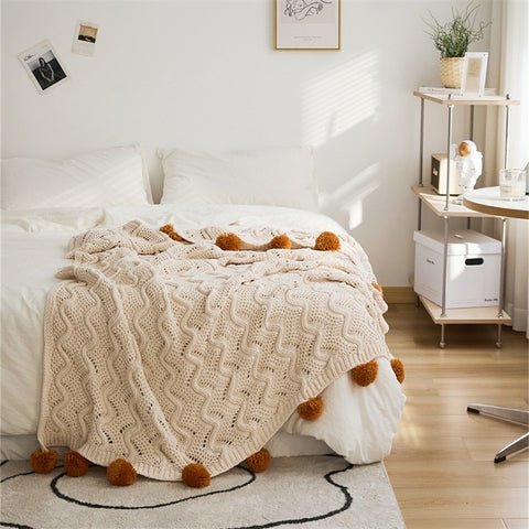 A gray Nordic-inspired knitted throw with a cable knit pattern and pom-pom details, draped over the arm of a beige armchair.