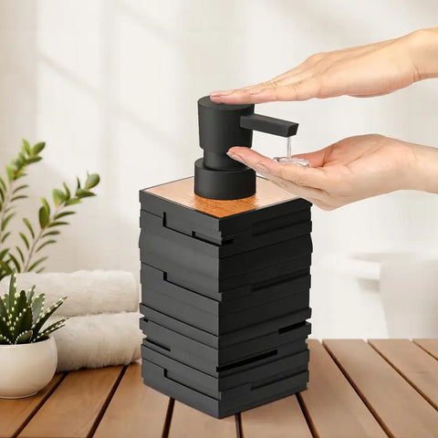 Black soap dispenser with wooden top on a wooden surface, hands reaching for it.