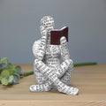 Decorative figure made of printed paper sitting and reading a book on a wooden surface.