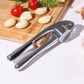 Garlic press on a white surface with garlic cloves and tomatoes on a wooden board in the background.