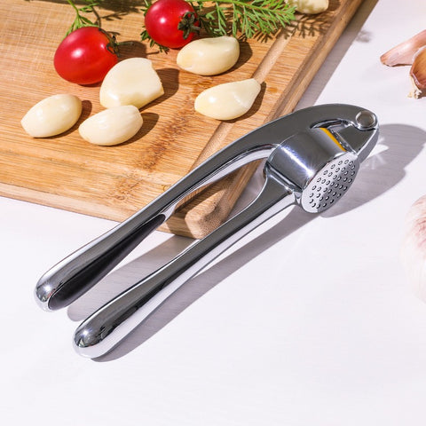 Garlic press on a white surface with garlic cloves and tomatoes on a wooden board in the background.