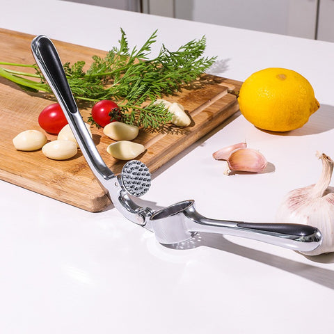 Garlic press on a kitchen counter with garlic cloves, lemon, and herbs.