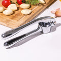 Garlic press on a white surface with garlic cloves and a cutting board in the background