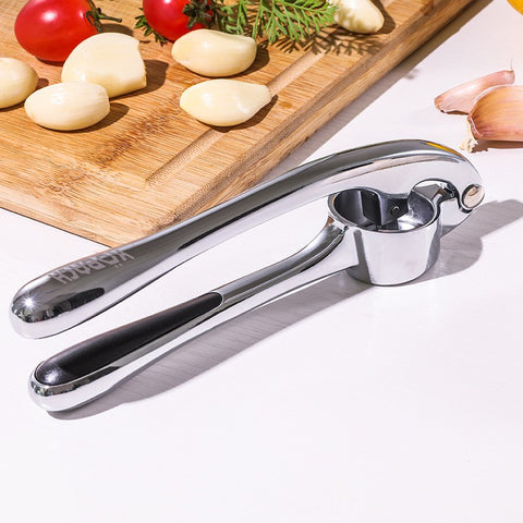 Garlic press on a white surface with garlic cloves and a cutting board in the background