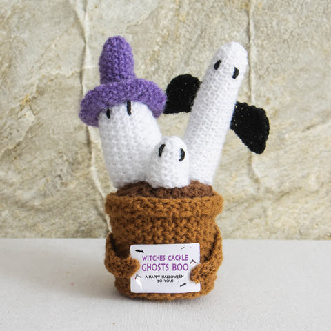 Crocheted Halloween-themed figurine with ghosts and a witch, held in a brown basket, against a stone wall background.
