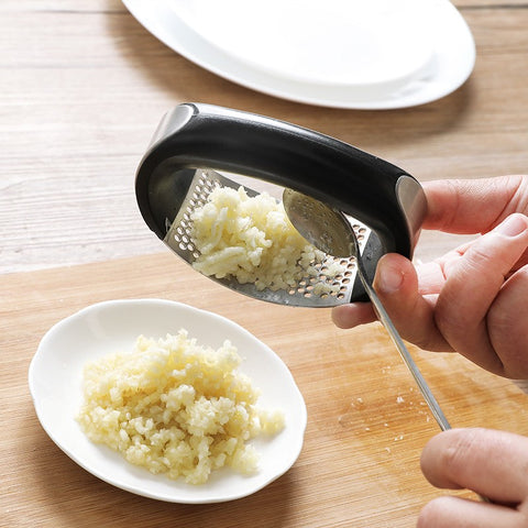 Hand using a garlic press to crush garlic on a wooden cutting board with a white plate in the background.