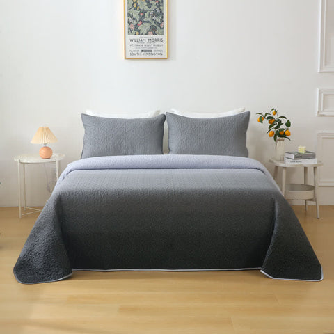 Gray quilt and pillows on a bed in a bedroom setting with a lamp and plant.