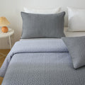 Gray quilted bedding set on a bed with white pillows and a side table with a lamp.