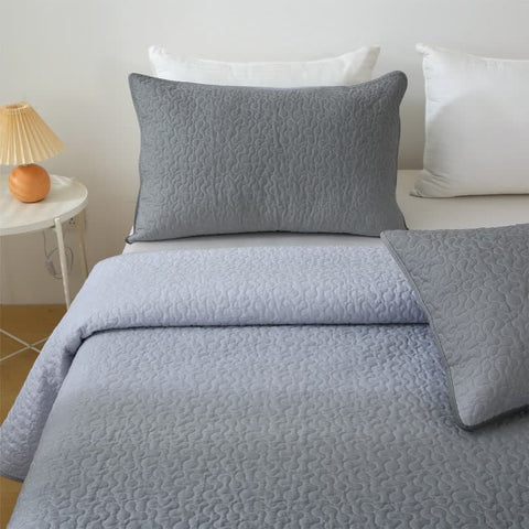 Gray quilted bedding set on a bed with white pillows and a side table with a lamp.