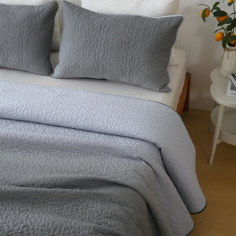 Gray textured quilt and pillows on a bed with a side table and decorative items in the background.