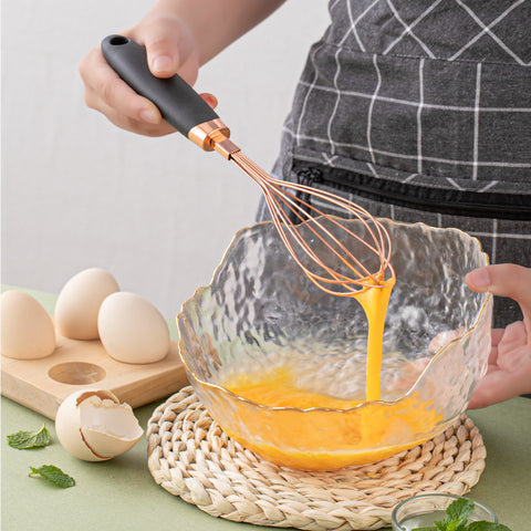 Person whisking fresh eggs in a glass bowl using a rose gold stainless steel whisk with black ergonomic handle. Durable balloon whisk perfect for baking, cooking, and everyday kitchen use.