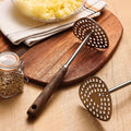 Metal potato masher on a wooden cutting board with a bowl of mashed potatoes and a jar of spices in the background.