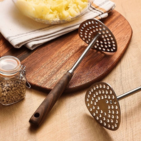 Metal potato masher on a wooden cutting board with a bowl of mashed potatoes and a jar of spices in the background.