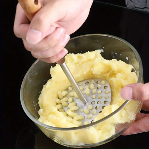 Mashed potatoes being transferred from a metal masher into a glass bowl.