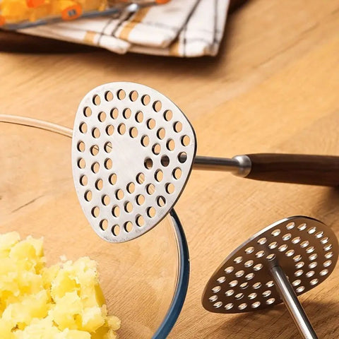 Metal potato masher on a wooden surface with mashed potatoes and a kitchen towel in the background.