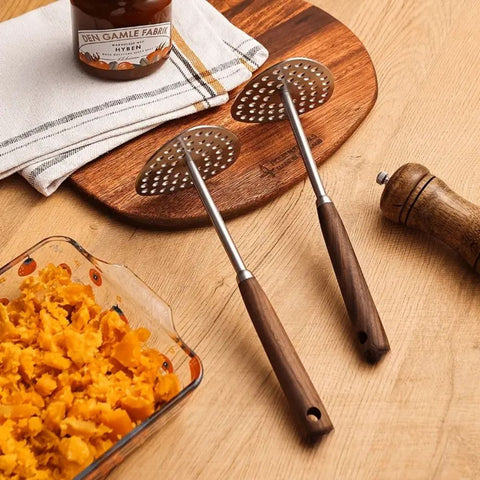 Two metal potato mashers with wooden handles on a wooden surface next to a dish of food and a bottle.