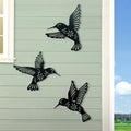 Three black metal hummingbird sculptures on a light green wall with a blue sky and trees in the background.