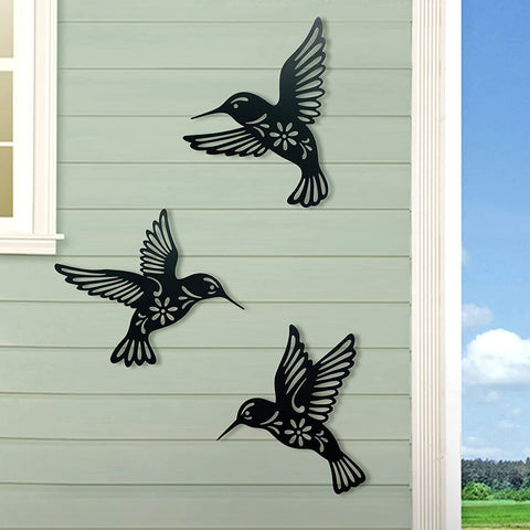 Three black metal hummingbird sculptures on a light green wall with a blue sky and trees in the background.