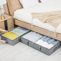 Gray storage bins under a bed with folded clothes inside, in a bedroom setting.