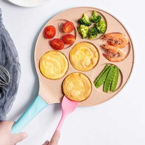 Animals waffle and pancake pan with blue handle cooking pancakes and assorted vegetables, hand holding pan and pink spatula on white surface
