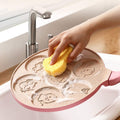Hand cleaning pink Animals Waffle & Pancake Pan with sponge, showing embossed animal shapes on non-stick surface