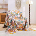 Autumn Garden Cotton Throw Blanket with warm floral patterns draped over a beige armchair in a cozy, bright living room setting