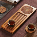 A bamboo tea tray with two cups of tea and coasters on a wooden surface.