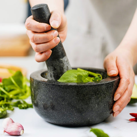 Hand holding black marble mortar pestle crushing green leaves with garlic and chili nearby