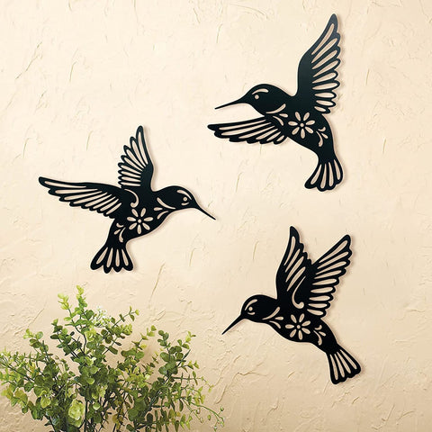 Three black metal hummingbird wall art pieces on a beige wall with a plant in the corner.