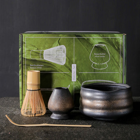 Tea-making set with black pot, whisk, and scoop in front of a green box on a dark surface.