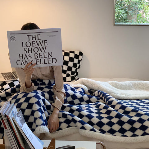 Person sitting on a couch with a checkered blanket, holding a newspaper with 'The Loewe Show Has Been Cancelled' headline.