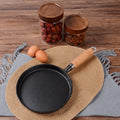 Black frying pan with wooden handle on a woven mat, eggs and jars in the background.