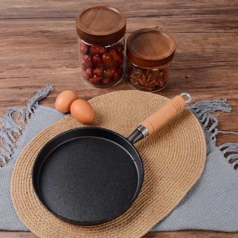 Black frying pan with wooden handle on a woven mat, eggs and jars in the background.