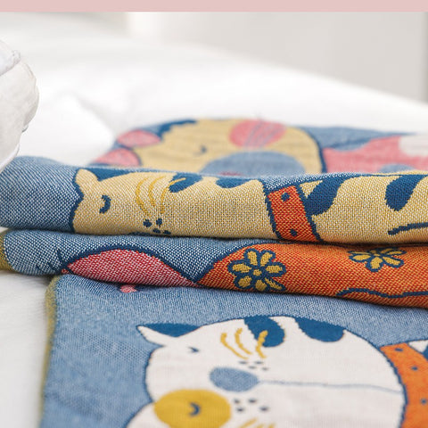 Close-up of Cat Party cotton pillowcase fabric showing detailed colorful cat illustrations on soft blue cotton material, folded on a white bed.