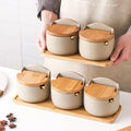 Set of ceramic containers with wooden lids on a wooden tray held by a person.