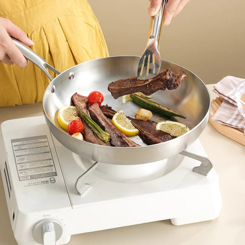 Person cooking steak and vegetables in stainless steel skillet on a portable stove.