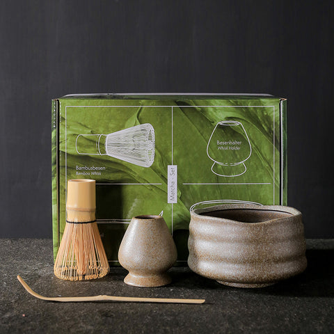 Tea-making set with ceramic bowl, whisk, and holder in front of a green box on a dark background